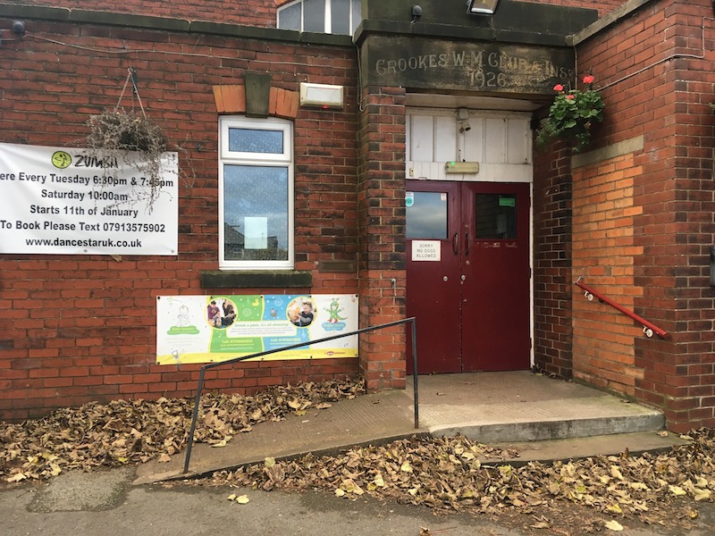 The front door of the venue, red double doors with a small concrete ramp and a handrail next to two steps and another handrail up to the door